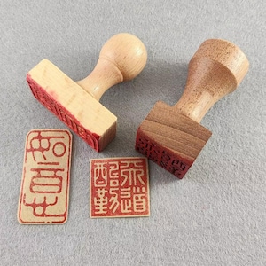 Puede incluir: Dos sellos de madera con impresiones de tinta roja y dos impresiones separadas de tinta roja. Los sellos tienen un mango de madera clara y una base rectangular o cuadrada. Las impresiones presentan caracteres asiáticos.