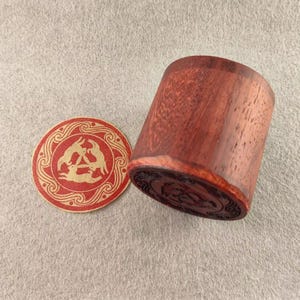 以下が含まれることがあります： 詳細なベースを備えた円筒形の赤茶色の木製容器と、丸いコースター。 コースターは、三角形の3匹のウサギの金色のデザインの赤い縁が特徴です。