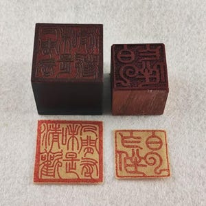 Puede incluir: Dos sellos chinos de madera con caracteres tallados. Un sello es de color marrón oscuro, el otro es de color marrón rojizo. También son visibles dos impresiones estampadas, que muestran los diseños de tinta roja sobre un fondo beige. Los sellos son cuadrados y las impresiones rectangulares.