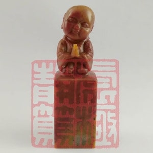 May include: A small, carved stone sculpture of a seated figure with a rounded face and hands clasped in prayer. The figure is wearing a robe and is sitting on a square base. The sculpture is made of a reddish-brown stone.