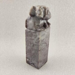 May include: A grey and white stone sculpture of a mythical creature, possibly a lion or a dragon, with a square base. The sculpture is carved in a detailed style and has a textured surface.