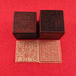 Puede incluir: Dos sellos de madera con intrincados diseños tallados, uno cuadrado y otro rectangular, se asientan sobre dos impresiones rojas sobre una superficie roja. Los sellos e impresiones presentan caracteres estilizados detallados.