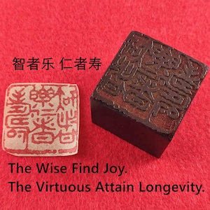Puede incluir: Un sello rectangular marrón oscuro con caracteres tallados y una imagen estampada más pequeña con tinta roja sobre un fondo beige. El texto dice: "The Wise Find Joy. The Virtuous Attain Longevity."