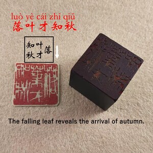 Puede incluir: Una piedra de tinta rectangular marrón oscuro con caracteres tallados y un sello de tinta roja con un diseño blanco. El texto "The falling leaf reveals the arrival of autumn" está debajo.
