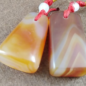 Puede incluir: Dos colgantes de piedra de ágata naranja y blanca con cordones rojos. Las piedras son rectangulares y tienen un patrón de remolino.