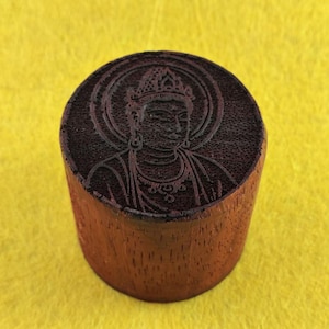 Puede incluir: Un sello cilíndrico de madera con un tallado detallado de una figura serena, probablemente un Buda o una deidad, en su superficie superior. El sello es de madera marrón rojiza y se encuentra sobre un fondo amarillo.