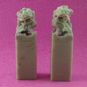 Puede incluir: Dos pequeñas esculturas de piedra talladas de leones, cada una sobre una base rectangular. Las esculturas son de color verde claro y tienen detalles intrincados.