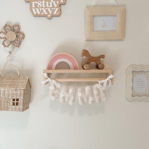 May include: A wooden wall shelf holding a pink and white rainbow and a wooden horse toy. Below the shelf hangs a fabric banner spelling EMMA in cream-coloured letters decorated with small white flowers, tied with white ribbons at each end.