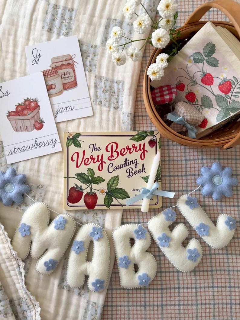 Puede incluir: Una naturaleza muerta encantadora con un libro infantil titulado "The Very Berry Counting Book" con ilustraciones de fresas. Una pancarta de fieltro deletrea "MABEL" con detalles de flores azules. Una cesta contiene servilletas y tarros de mermelada.