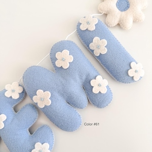 Puede incluir: Un bander&iacute;n de fieltro azul claro que deletrea la palabra "HOME", decorado con flores de fieltro blanco. El bander&iacute;n est&aacute; ensartado en una cuerda blanca, con una gran flor blanca al final. Es un art&iacute;culo decorativo.