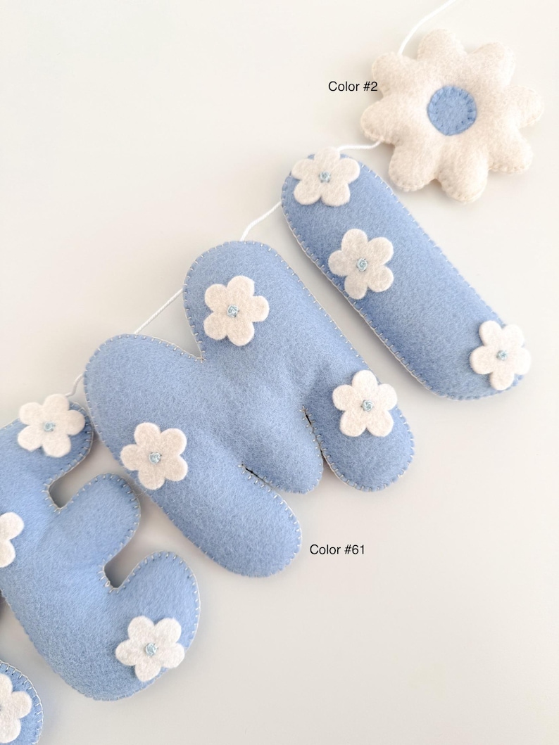 Puede incluir: Un bander&iacute;n de fieltro azul claro que deletrea la palabra "HOME", decorado con flores de fieltro blanco. El bander&iacute;n est&aacute; ensartado en una cuerda blanca, con una gran flor blanca al final. Es un art&iacute;culo decorativo.