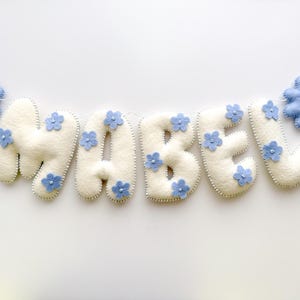 Puede incluir: Un cartel de fieltro crema con el nombre "MABEL", adornado con peque&ntilde;as flores azules. Dos flores azules m&aacute;s grandes flanquean el nombre. El cartel est&aacute; ensartado en un cord&oacute;n blanco, perfecto para la decoraci&oacute;n de la guarder&iacute;a.