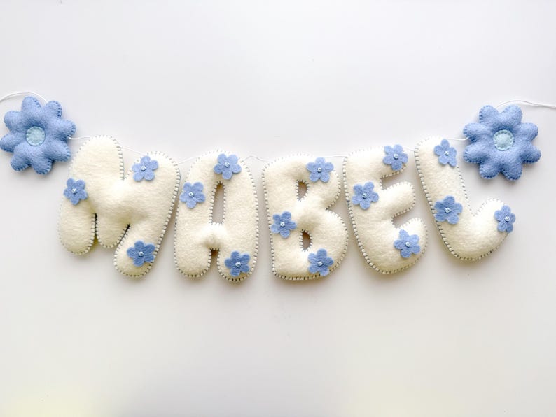 Puede incluir: Un cartel de fieltro crema con el nombre "MABEL", adornado con peque&ntilde;as flores azules. Dos flores azules m&aacute;s grandes flanquean el nombre. El cartel est&aacute; ensartado en un cord&oacute;n blanco, perfecto para la decoraci&oacute;n de la guarder&iacute;a.