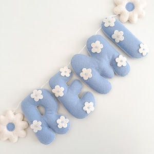 Puede incluir: Un cartel de fieltro azul claro que deletrea el nombre "REMY" con detalles de flores blancas. Las letras est&aacute;n conectadas por una cuerda blanca, y el cartel incluye detalles de flores de fieltro blanco en cada extremo. Sobre un fondo blanco.