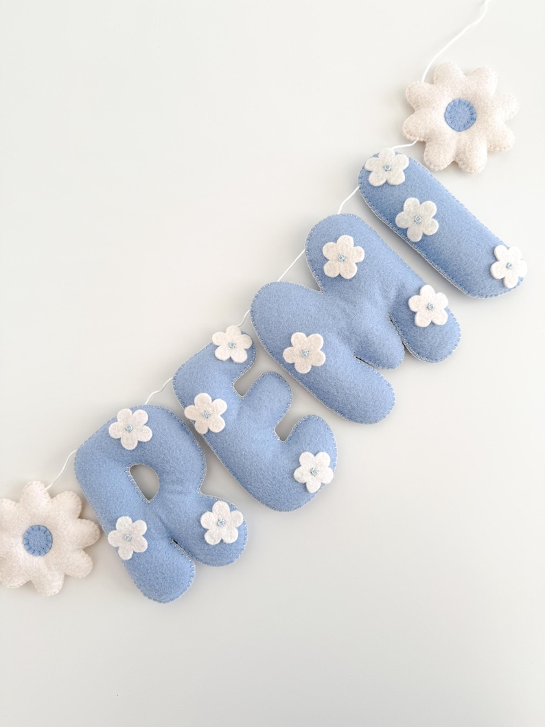 Puede incluir: Un cartel de fieltro azul claro que deletrea el nombre "REMY" con detalles de flores blancas. Las letras est&aacute;n conectadas por una cuerda blanca, y el cartel incluye detalles de flores de fieltro blanco en cada extremo. Sobre un fondo blanco.