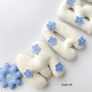 Puede incluir: Un bander&iacute;n de fieltro crema con la palabra "BABY", adornado con flores de fieltro azul claro. Las letras est&aacute;n delineadas con costuras grises, y una gran flor azul est&aacute; a la izquierda. El texto "Color #4" est&aacute; en la parte inferior derecha.