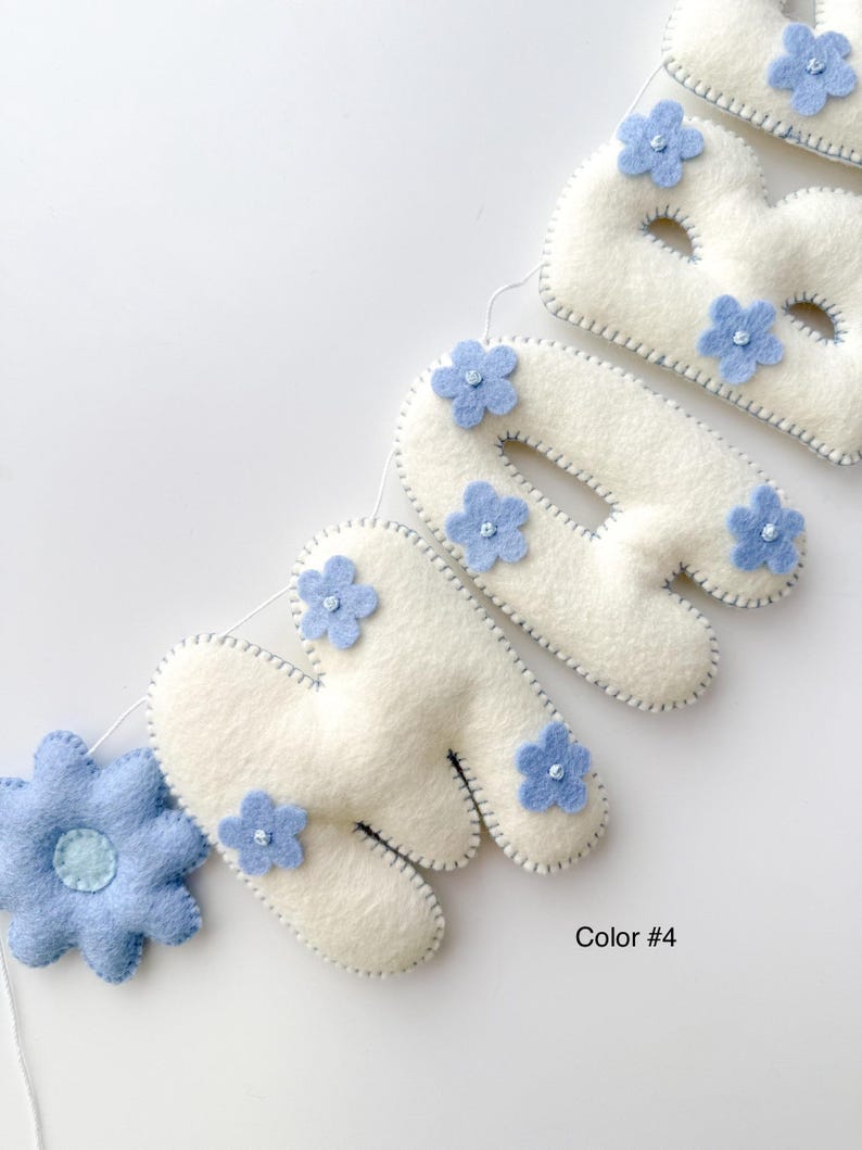 Puede incluir: Un bander&iacute;n de fieltro crema con la palabra "BABY", adornado con flores de fieltro azul claro. Las letras est&aacute;n delineadas con costuras grises, y una gran flor azul est&aacute; a la izquierda. El texto "Color #4" est&aacute; en la parte inferior derecha.