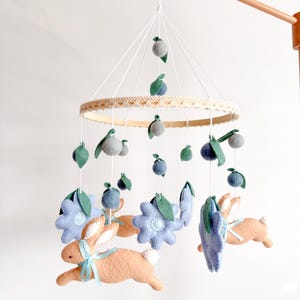 Puede incluir: Móvil para bebé con conejitos de fieltro, flores azules y formas grises de bayas, suspendido de un marco de madera. El móvil tiene un anillo ribeteado de encaje color crema, ideal para una habitación infantil.