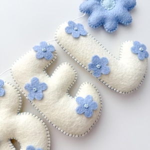 Puede incluir: Un cartel de fieltro que deletrea la palabra "BABY" en letras blancas, cada una adornada con flores de fieltro azul claro. Las letras est&aacute;n cosidas con un hilo azul contrastante, y el cartel est&aacute; ensartado en un cord&oacute;n blanco. Tambi&eacute;n se incluye una flor azul grande.