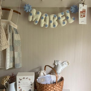 Puede incluir: Un cartel de fieltro con la palabra "MABEL" con detalles de flores azules, colgado sobre una cesta de mimbre y un libro decorativo. El cartel est&aacute; sujeto a una pared con peque&ntilde;as clavijas blancas.