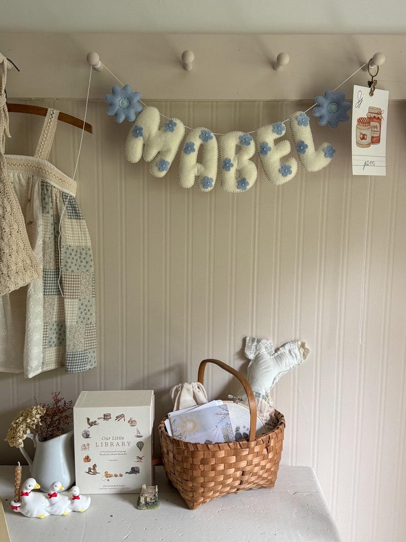 Puede incluir: Un cartel de fieltro con la palabra "MABEL" con detalles de flores azules, colgado sobre una cesta de mimbre y un libro decorativo. El cartel est&aacute; sujeto a una pared con peque&ntilde;as clavijas blancas.