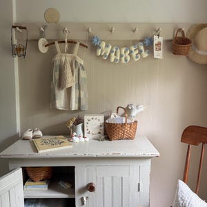 Puede incluir: Un acogedor cuarto de ni&ntilde;os con un armario blanco, una silla de madera y un perchero de pared. Una pancarta con la palabra "MABEL" cuelga sobre un vestido y una peque&ntilde;a cesta. Un libro y zapatos est&aacute;n sobre el armario.