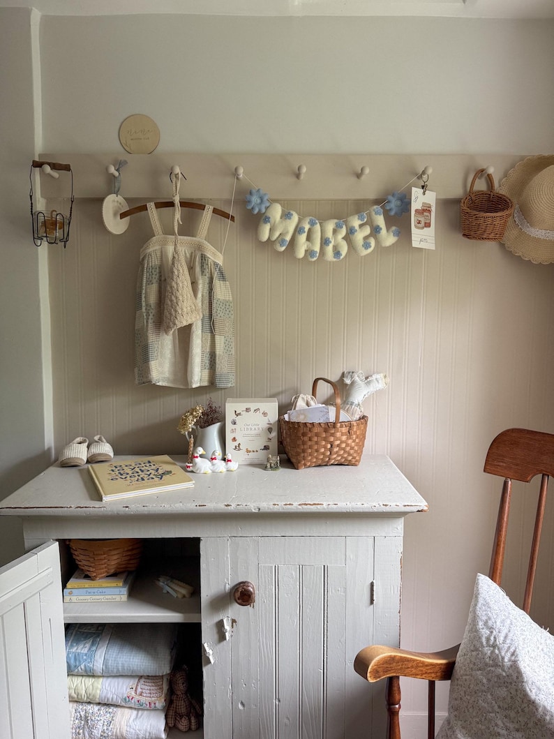 Puede incluir: Un acogedor cuarto de ni&ntilde;os con un armario blanco, una silla de madera y un perchero de pared. Una pancarta con la palabra "MABEL" cuelga sobre un vestido y una peque&ntilde;a cesta. Un libro y zapatos est&aacute;n sobre el armario.