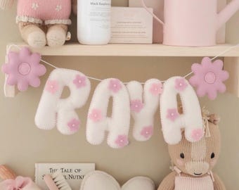 May include: A nursery shelf displays a felt banner spelling "RAYA" with pink flower accents. The shelf also holds a plush horse, a heart-shaped pillow, a doll, a watering can, and a bottle of lotion. The color palette is soft pink and white.