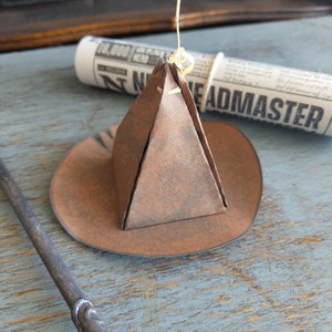 May include: A brown paper origami witch hat with a wide brim, suspended by a string. A rolled-up newspaper with the word "HEADMASTER" is in the background, along with a wand.