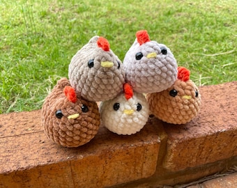 May include: A collection of six plush, crocheted chicken toys in various shades of brown, gray, and white. Each chicken has black eyes, a yellow beak, and a red comb. The toys are stacked on a brick surface with a green background.