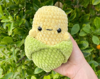 May include: A plush toy shaped like an ear of corn, featuring a textured yellow top with two small black bead eyes and a thin black stitched mouth, nestled inside a soft, light green crocheted husk that wraps around the base of the corn.