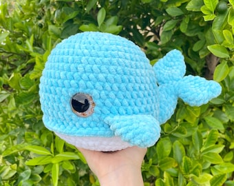 May include: A light blue crocheted whale plush toy with a white belly and brown eye. The whale has a textured, soft appearance and is held against a backdrop of green foliage. The plush toy is a handmade item.
