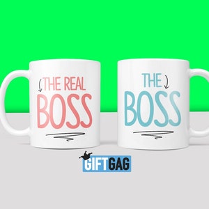 Puede incluir: Dos tazas de cer&aacute;mica blanca con texto negro. La taza de la izquierda dice "The Real Boss" en texto rosa con una flecha apuntando al texto. La taza de la derecha dice "The Boss" en texto azul con una flecha apuntando hacia afuera del texto.