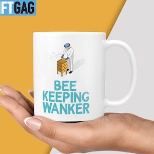 May include: White ceramic mug featuring a blue and white illustration of a beekeeper and a beehive. The text "BEE KEEPING WANKER" is printed in blue on the mug.