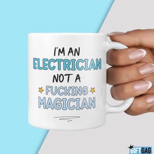 May include: White ceramic mug with the text "I'M AN ELECTRICIAN NOT A FUCKING MAGICIAN" in blue and black lettering. The mug has a white handle and is held by a person.