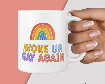 May include: White ceramic mug with a rainbow design and the text "WOKE UP GAY AGAIN" in colorful letters. The mug has a curved handle and is held by a hand with manicured nails. The mug is a fun and colorful item.