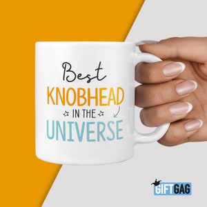 May include: White ceramic mug with the text "Best Knobhead in the Universe" in orange and blue lettering.