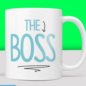 Puede incluir: Taza de cer&aacute;mica blanca con un dise&ntilde;o azul y negro. La taza dice "THE BOSS" con una flecha apuntando hacia abajo.