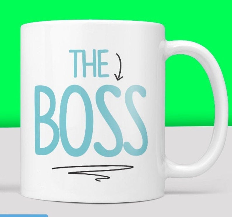 Puede incluir: Taza de cer&aacute;mica blanca con un dise&ntilde;o azul y negro. La taza dice "THE BOSS" con una flecha apuntando hacia abajo.