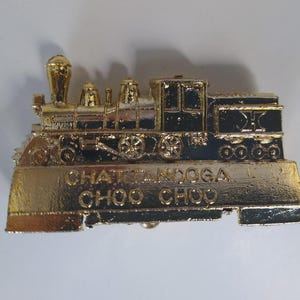 May include: A gold-toned metal belt buckle shaped like a steam locomotive train. The design features black enamel detailing on the engine and wheels. The text CHATTANOOGA CHOO CHOO is embossed in gold lettering on the rectangular base of the buckle.