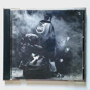 May include: A black and white CD case for The Who's Quadrophenia album. The cover features a person standing next to a scooter, wearing a jacket with the word WHO on the back. The text QUADROPHENIA is printed vertically along the sides.