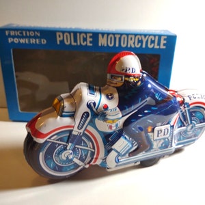 May include: A blue and white friction-powered toy police motorcycle with a rider wearing a blue uniform and a red helmet with "P.D." written on it. The motorcycle has a yellow headlight and "P.D." written on the side.