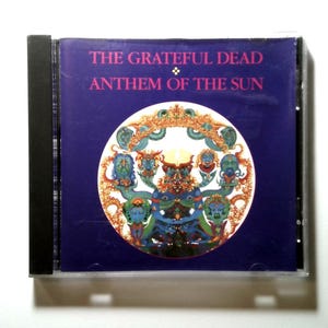 May include: A CD case for "Anthem of the Sun" by The Grateful Dead. The cover is dark blue with the band's name and album title in pink. A circular graphic features colorful, stylized figures.
