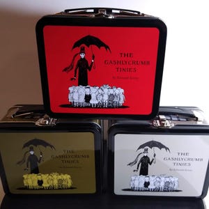 May include: Three metal lunchboxes with black frames and clasps. Each box has a different coloured front panel with the title "THE GASHLYCRUMB TINIES" and an illustration of a figure with an umbrella and a crowd of children.