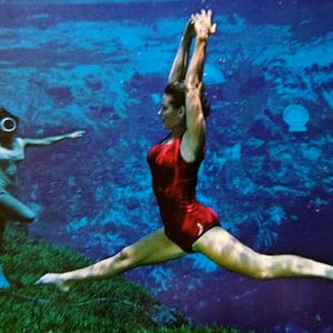 Könnte beinhalten: Unterwasserszene mit zwei Personen. Eine Person in einem roten Badeanzug macht einen Spagat mit erhobenen Armen. Die andere, in heller Kleidung, scheint einen Taucherhelm zu tragen. Der Hintergrund ist eine blaue Unterwasserumgebung.