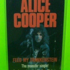 May include: A black and red Alice Cooper promotional card in a clear plastic holder. The card features the text "ALICE COOPER" in large red letters above an image of the artist. Below the image, the text reads "FEED MY FRANKENSTEIN" and "WAYNE'S WORLD".