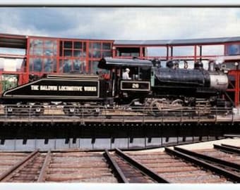 May include: A vintage black steam locomotive with the text "THE BALDWIN LOCOMOTIVE WORKS" on the side. The locomotive is on a turntable, with a red and glass building in the background. A person is visible in the cab.