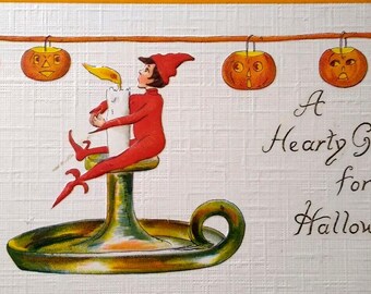May include: A vintage Halloween greeting card featuring a small elf sitting on a candlestick with a lit candle. The elf is wearing a red and white costume. There are three jack-o'-lanterns hanging from a string above the elf. The card reads "A Hearty Greeting for Halloween."