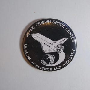 May include: A round, black button featuring a white illustration of a space shuttle and a globe. The button has the text "HENRY CROWN SPACE CENTER MUSEUM OF SCIENCE AND INDUSTRY" in white lettering.
