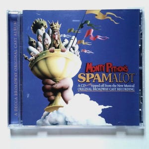 May include: A Monty Python's Spamalot original Broadway cast recording CD in a clear plastic jewel case. The cover features a large golden chalice held by a hand, with a king and knights inside, set against a deep blue background with clouds.